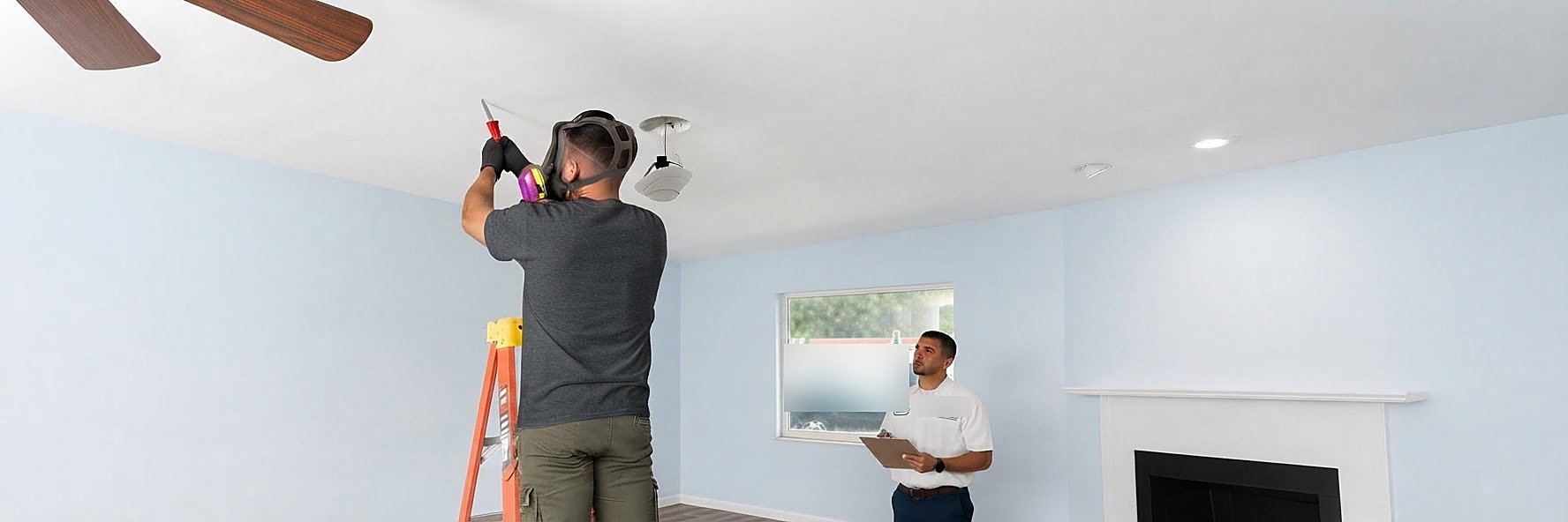Technician in a respirator repairing a ceiling while a supervisor reviews the job on a clipboard