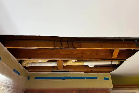 Opened ceiling cavity showing drain pipes and water-stained joists above a living space